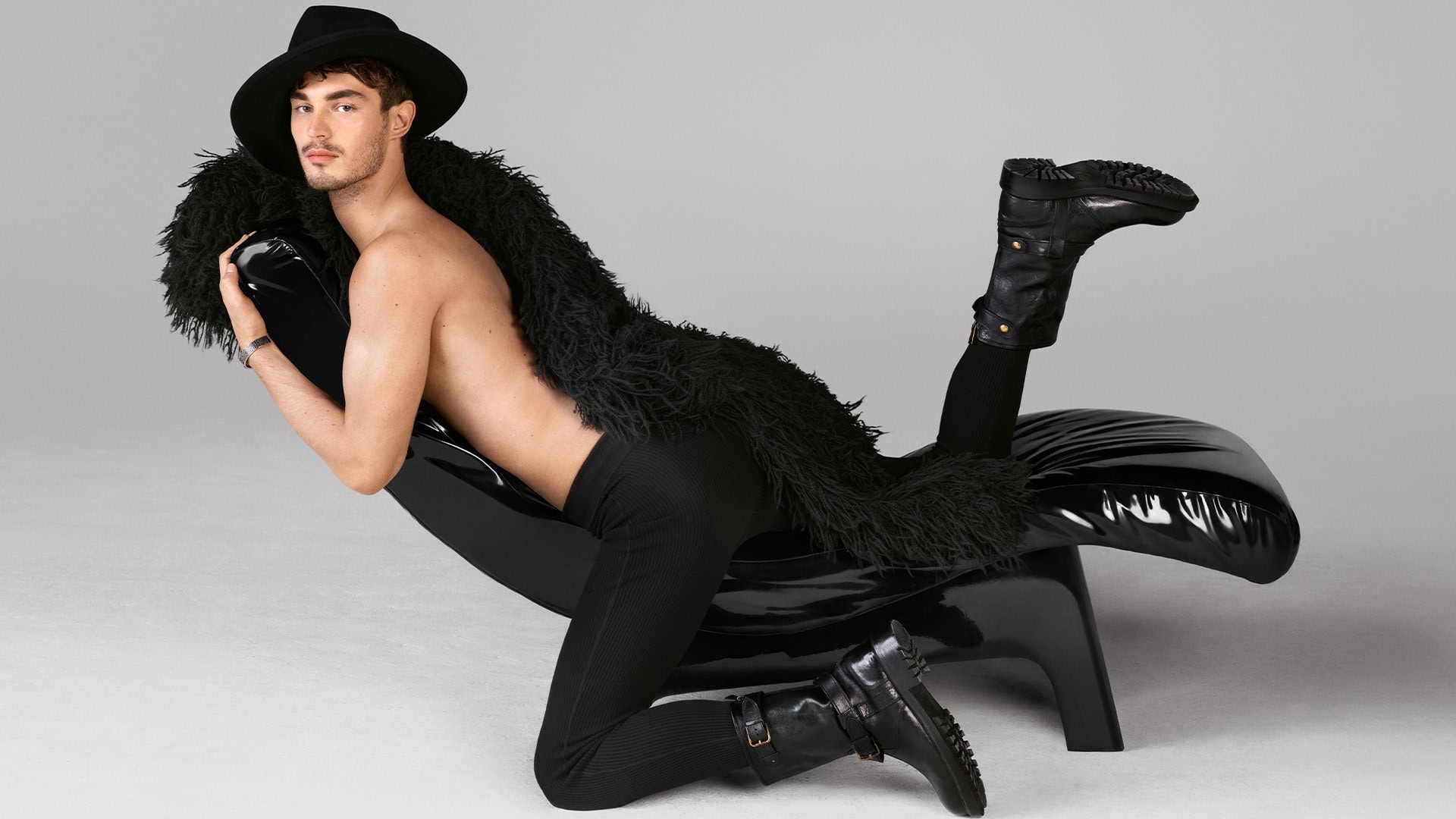 Adult male fashion model poses reclining on a black chair with a black fur coat, black hat, leggings, and boots on a grey background.