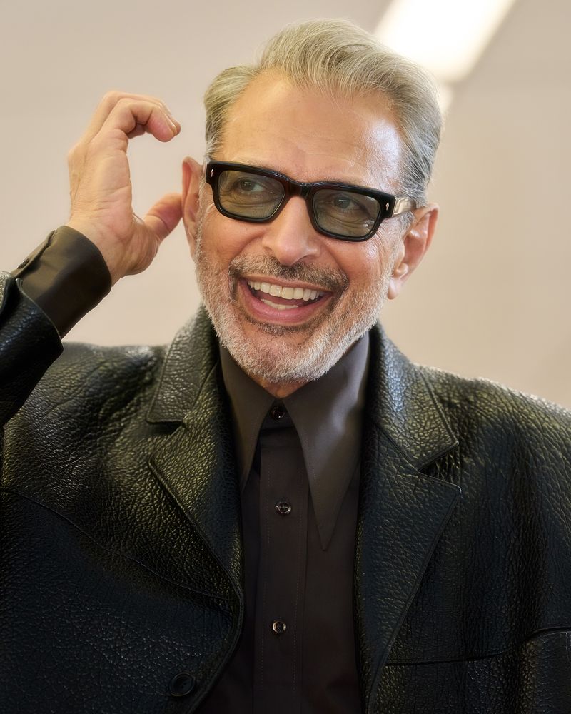 Jeff Goldblum smiles and poses in a stylish black leather jacket and dark glasses, indoors against a plain background in a medium shot.