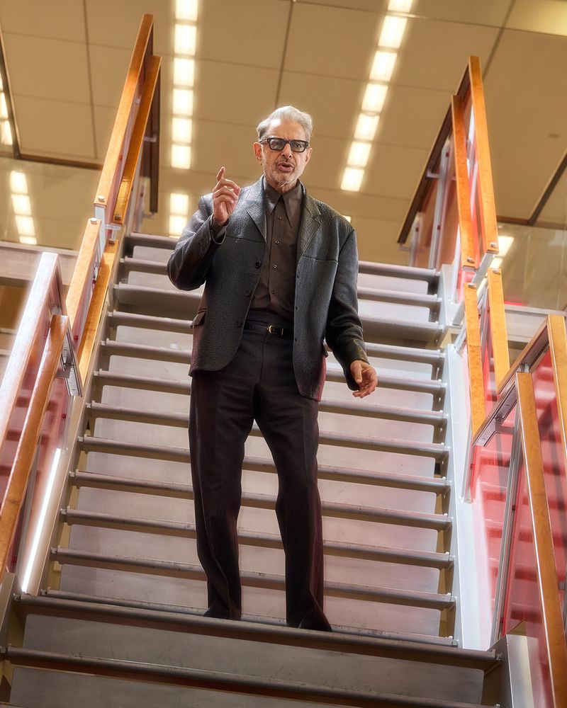 Adult male with glasses stands on staircase in a suit jacket and pants, facing forward, with his right hand raised in a building.