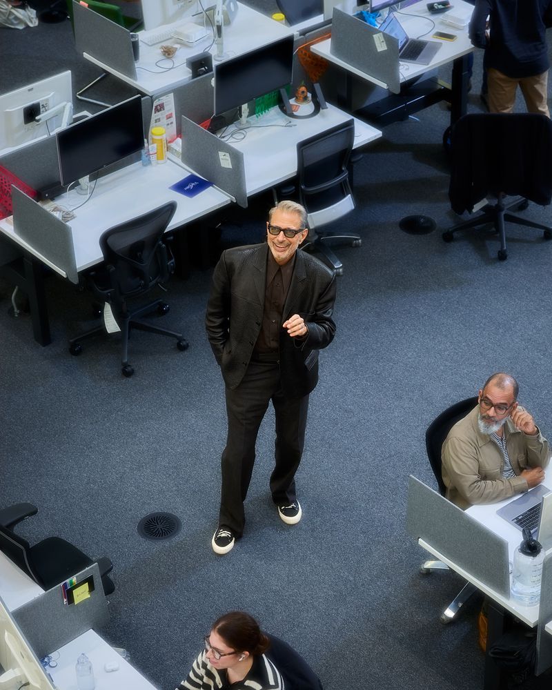 High-angle view of an adult male in a suit standing in an office surrounded by desks and computers, with additional office workers visible.