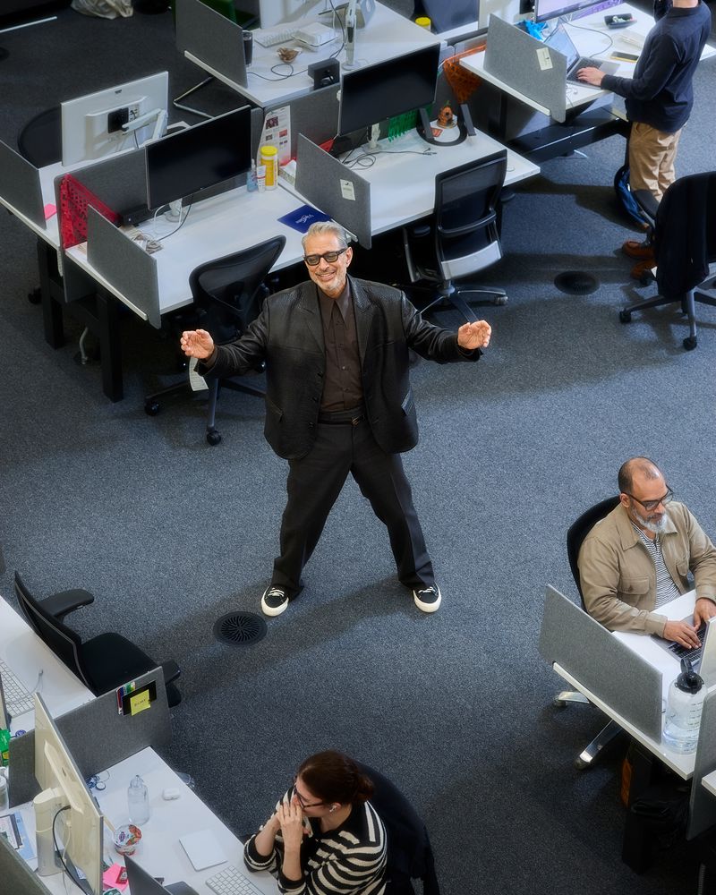 Overhead view captures a cheerful man in black blazer stands in an office setting with desks and coworkers, creating a lively workspace.