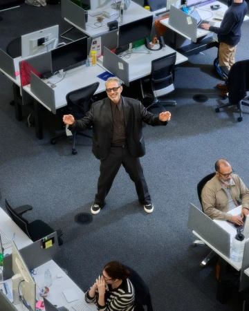 Overhead view captures a cheerful man in black blazer stands in an office setting with desks and coworkers, creating a lively workspace.