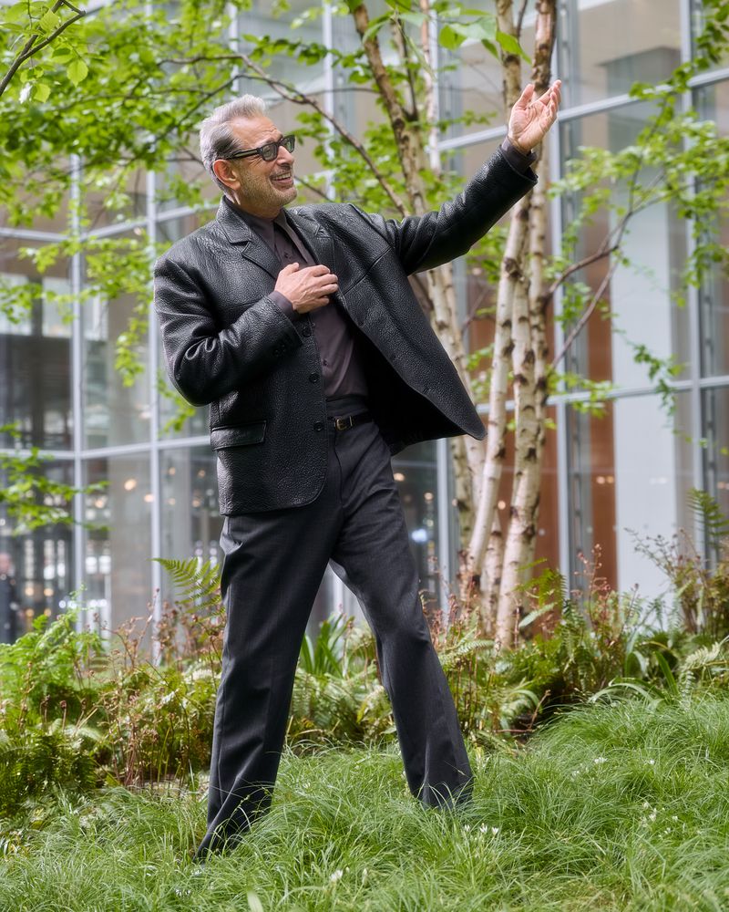 Jeff Goldblum stands in a park with trees and modern architecture, wearing a stylish dark suit and glasses, with a cheerful expression.