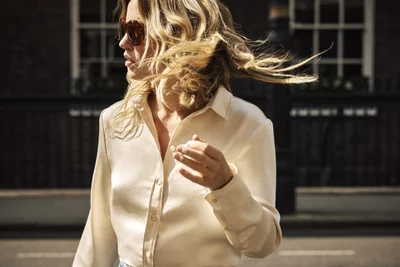 Georgia May Jagger wearing a cream silk button-down shirt and tortoiseshell sunglasses with windblown blonde hair on a city street.