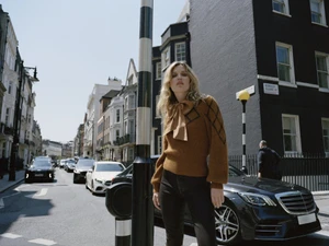 Georgia May Jagger wearing a brown pussy-bow sweater and dark jeans standing on a London street for 7 For All Mankind