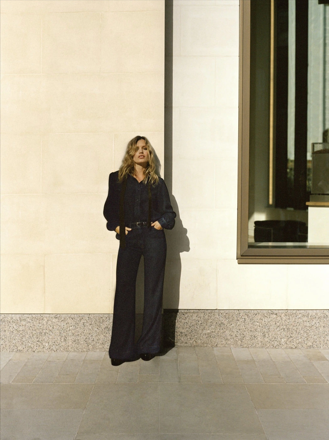 Georgia May Jagger wearing a dark denim jumpsuit standing against a light stone wall for 7 For All Mankind