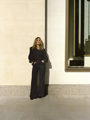 Georgia May Jagger wearing a dark denim jumpsuit standing against a light stone wall for 7 For All Mankind