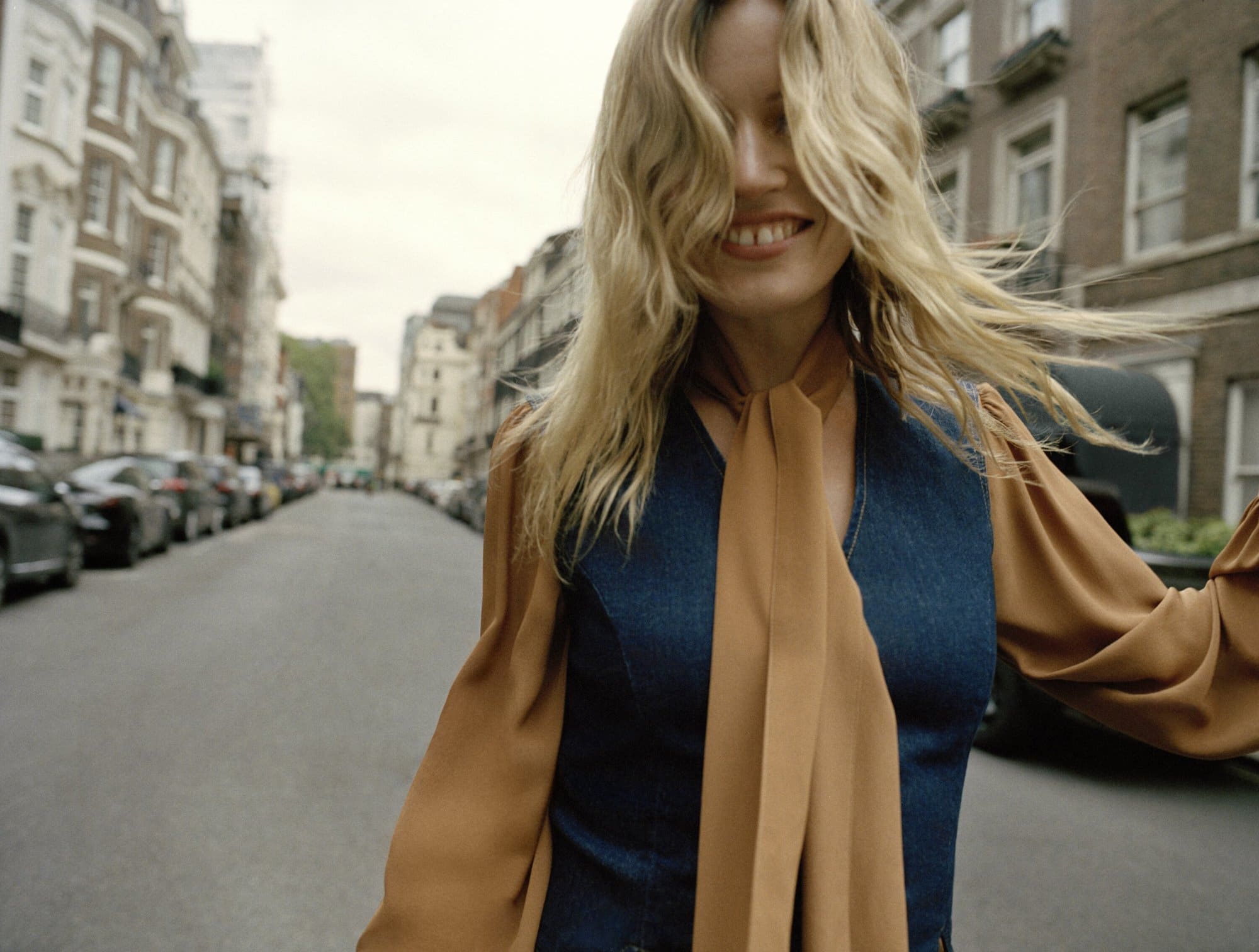 Smiling blonde woman in a blouse and vest walks down a city street. Buildings and parked cars line the sides of the road.