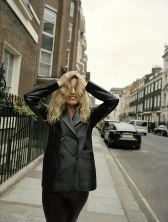 Blonde woman in black leather-like blazer poses on city street with residential buildings and cars. Hands on head, candid fashion shot.