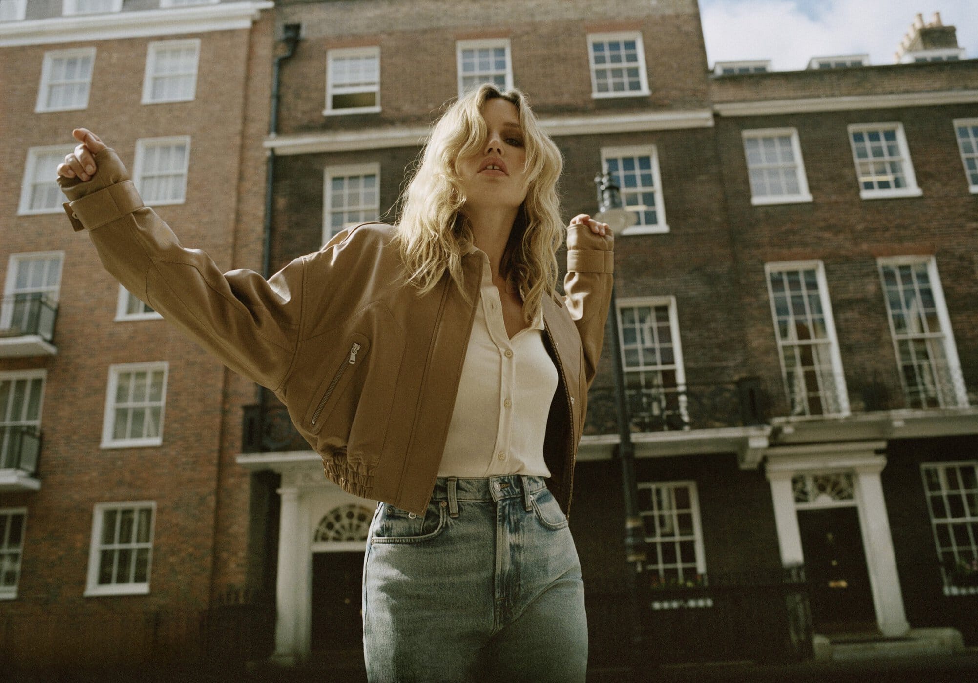 A blonde woman in a leather jacket stands outdoors with arms raised against an urban brick building, showcasing fashion and lifestyle photography.