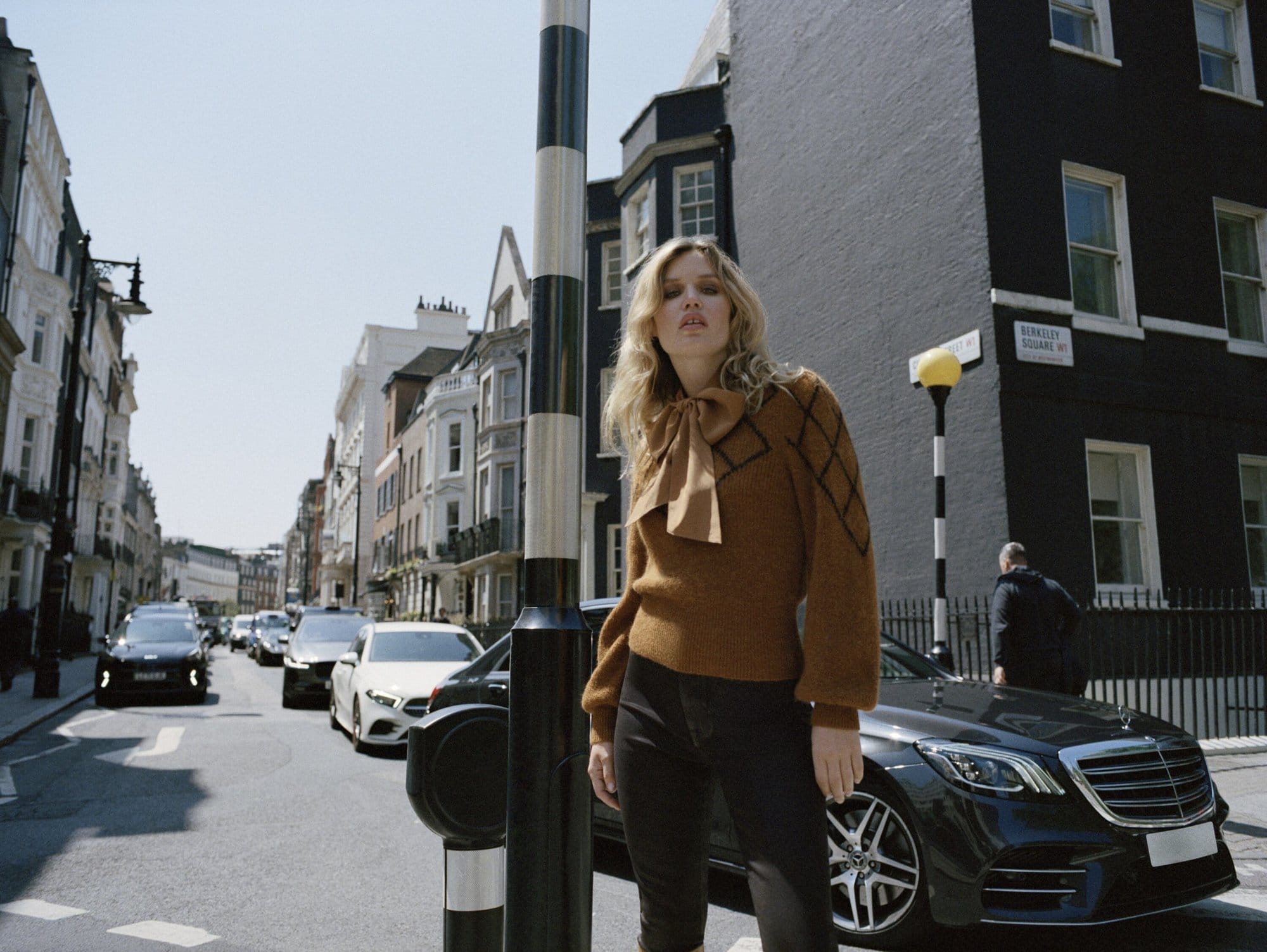 A woman poses in a city street wearing a brown sweater and black jeans, standing near a black car, with buildings in the background.