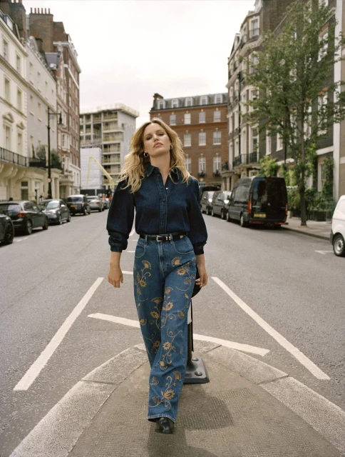 A woman in jeans and a denim shirt walks on an empty city street lined with parked cars and buildings on an overcast day.