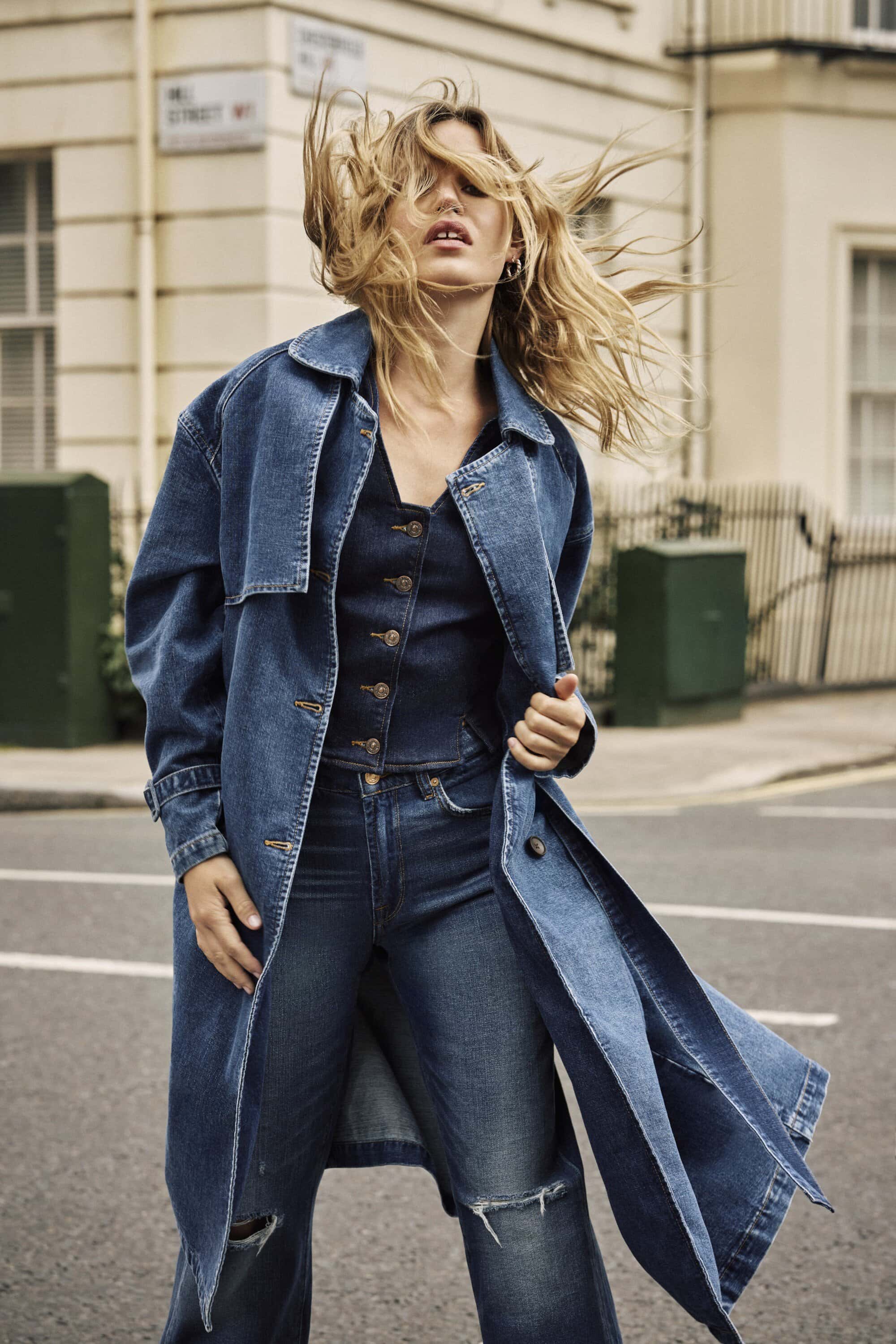 Woman in denim trench coat, jeans, and vest walking on a city street. Long blonde hair is tossed by the wind. Fashion photography.
