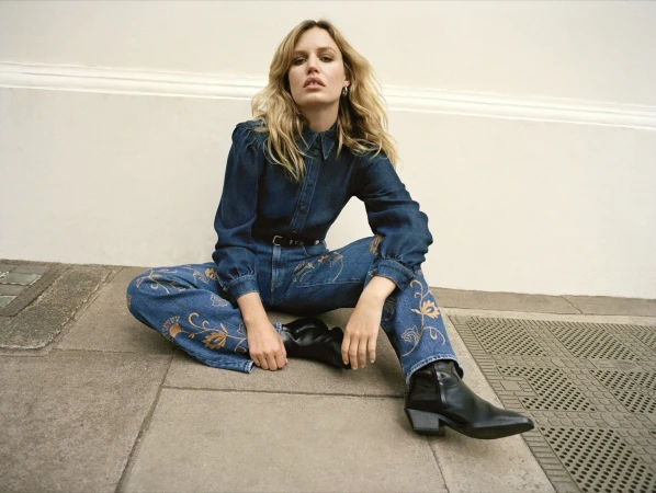 A woman sits in a cross-legged pose wearing a denim outfit with floral embroidery and black ankle boots, leaning against a wall outdoors.