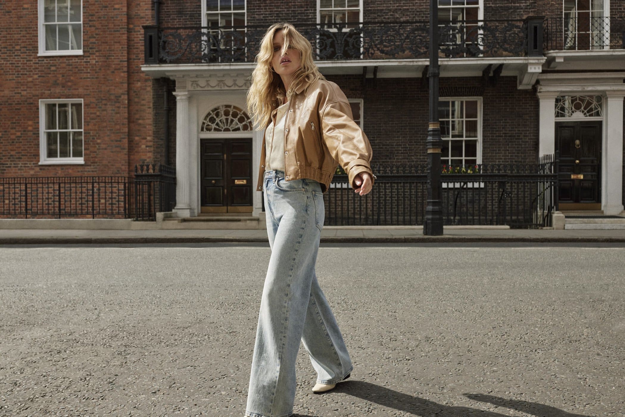 A blonde adult woman in a tan jacket and wide-leg blue jeans walks on a city street with brick buildings in the background.