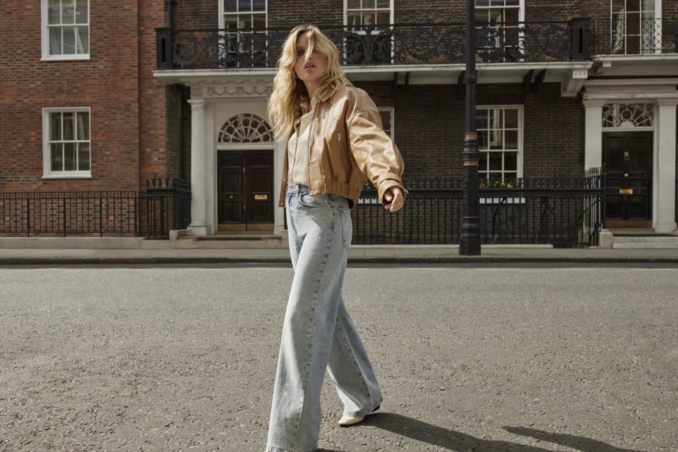 A blonde adult woman in a tan jacket and wide-leg blue jeans walks on a city street with brick buildings in the background.