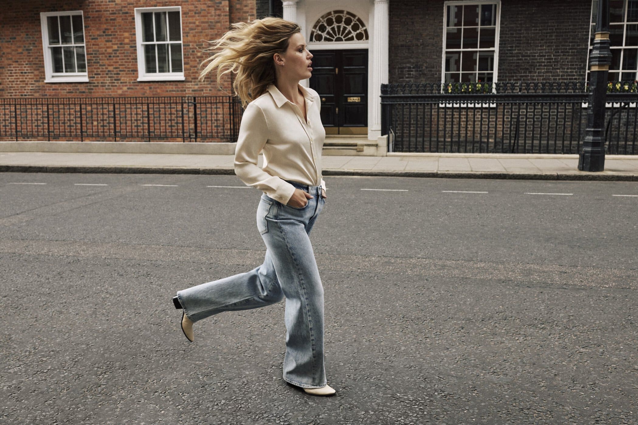 A woman in light-wash jeans and a cream blouse runs across an urban street in front of brick buildings with white-framed windows.