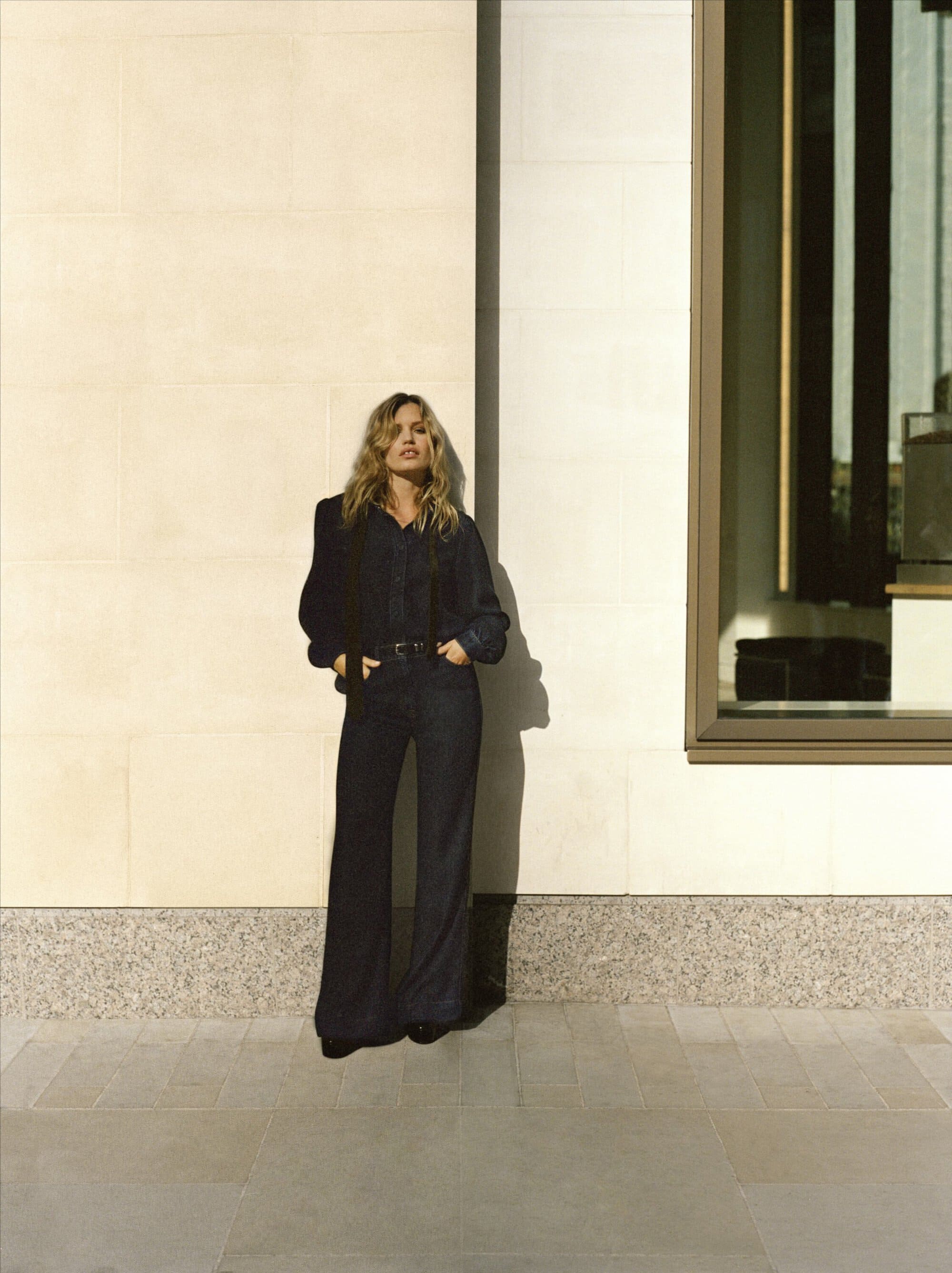 Adult woman in a denim outfit poses against a neutral wall with an architectural window reflection during daytime in an urban setting.