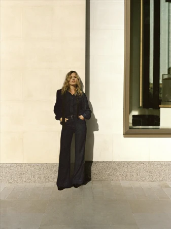 Adult woman in a denim outfit poses against a neutral wall with an architectural window reflection during daytime in an urban setting.