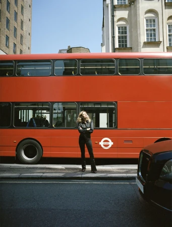 Woman poses in front of a red double-decker bus on a city street with tall buildings, wearing a black jacket and pants, standing in sunlight.