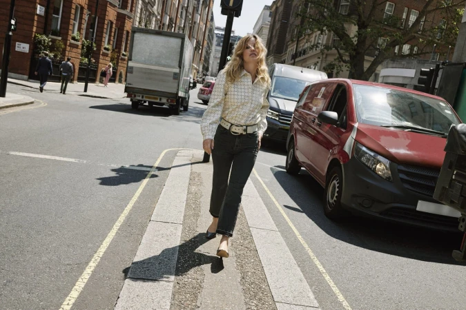 A blonde woman walks across a crosswalk on a city street, wearing a patterned blouse, dark jeans, and flats, with cars nearby.