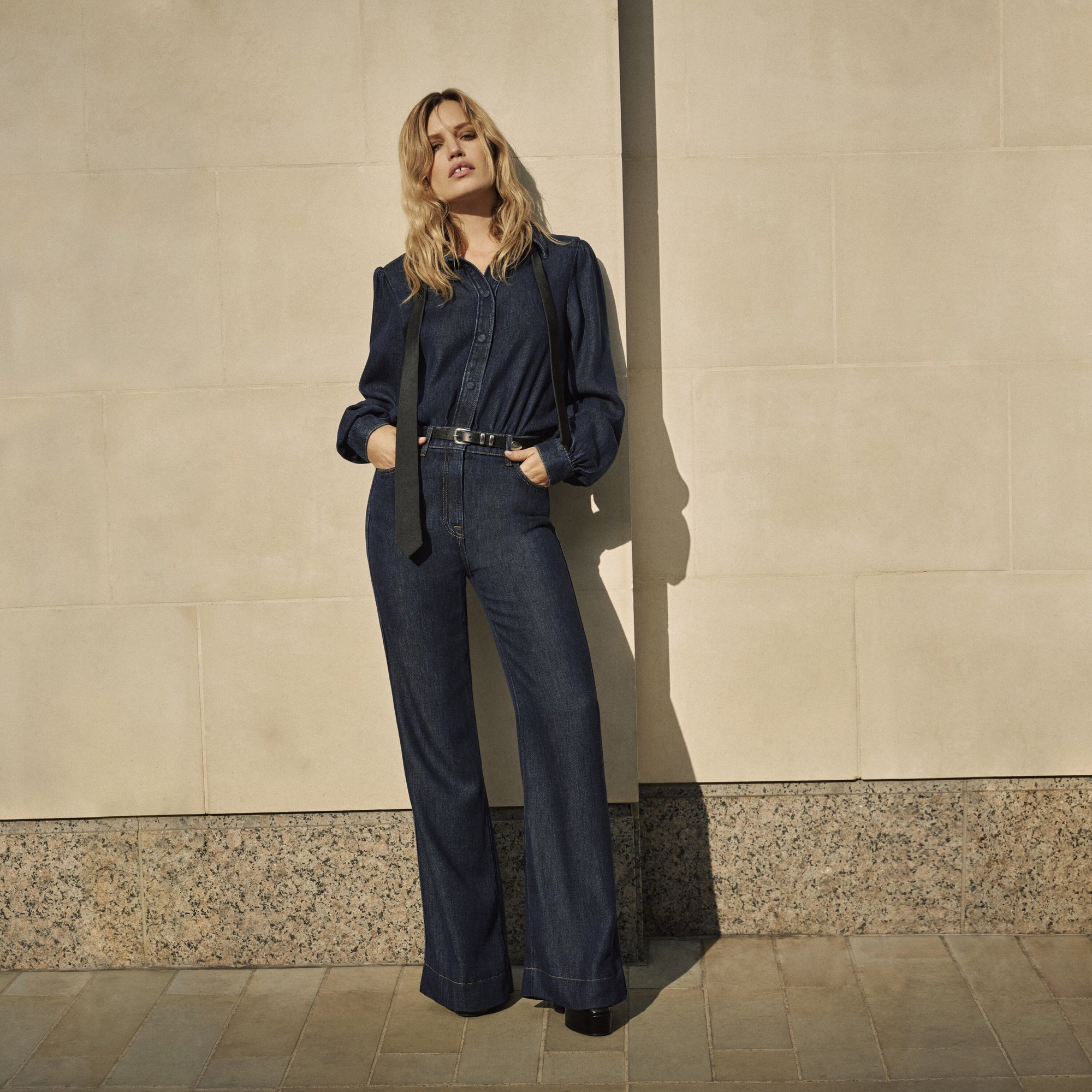 Fashionable woman in a dark denim jumpsuit with hands in her pockets stands against a neutral textured wall in direct sunlight.