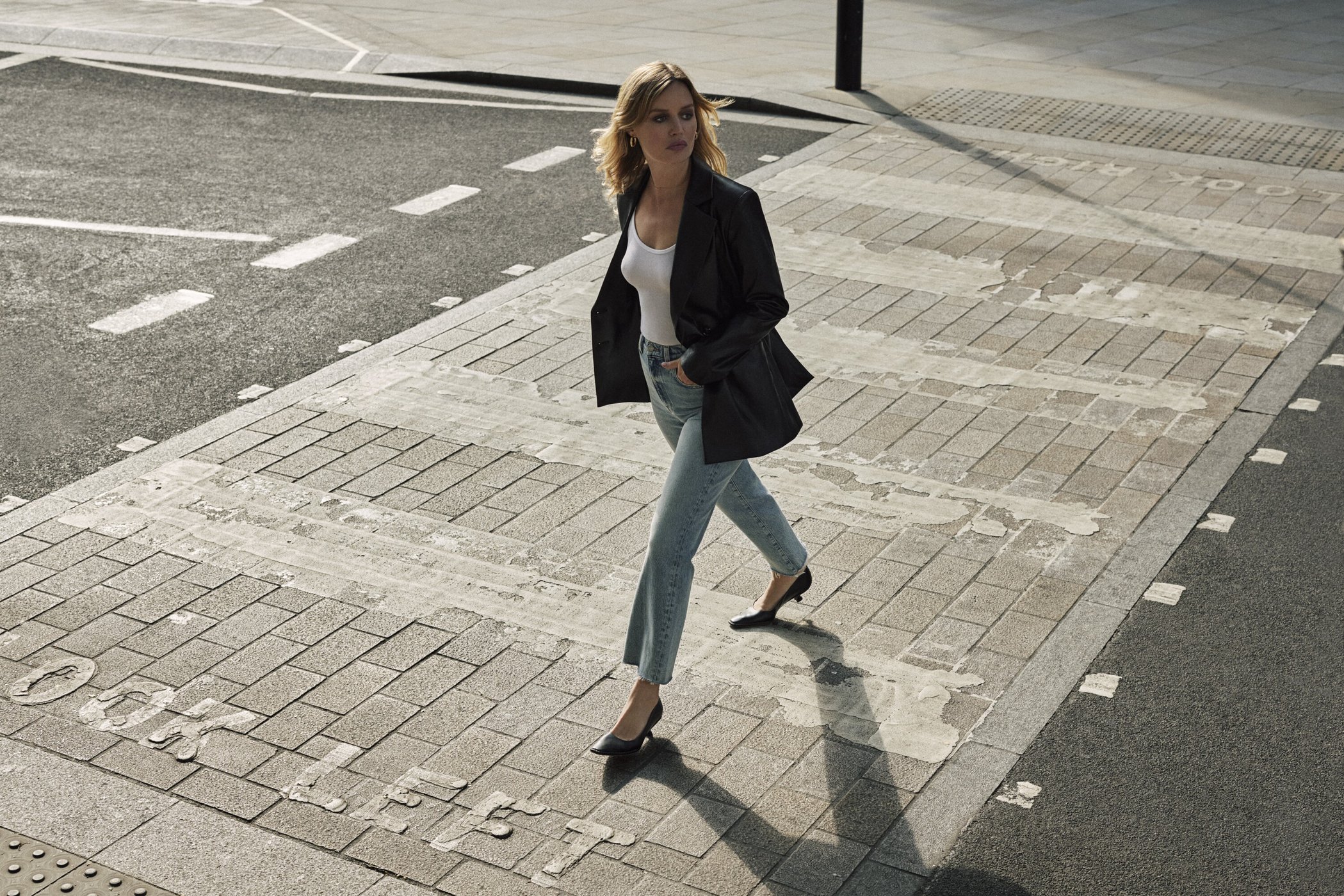 Adult female model walks across a city crosswalk in a black blazer, white top, light-wash jeans, and black heels in an urban setting.