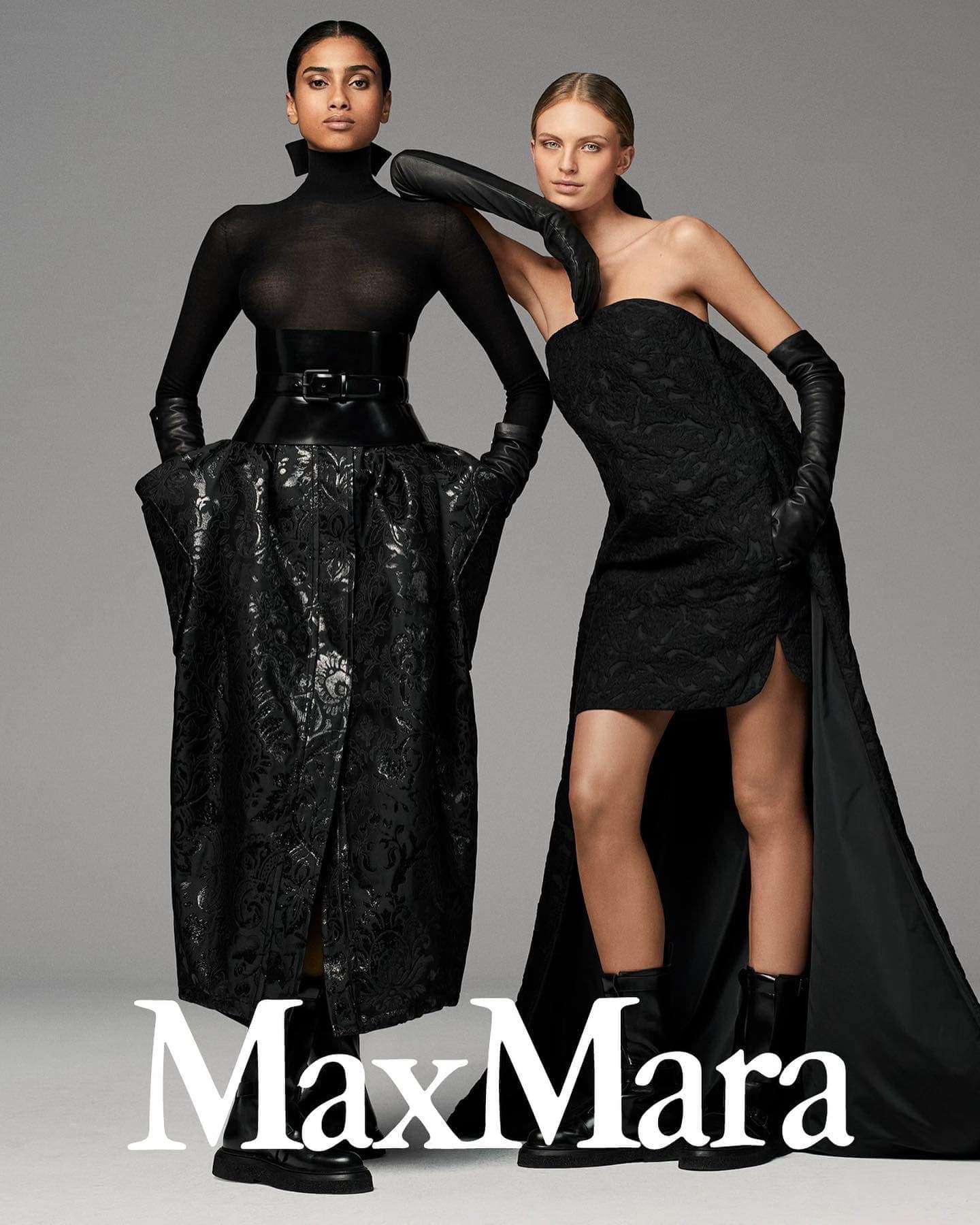 Two fashion models in black luxury outfits and opera gloves pose against a neutral grey studio background for a campaign.