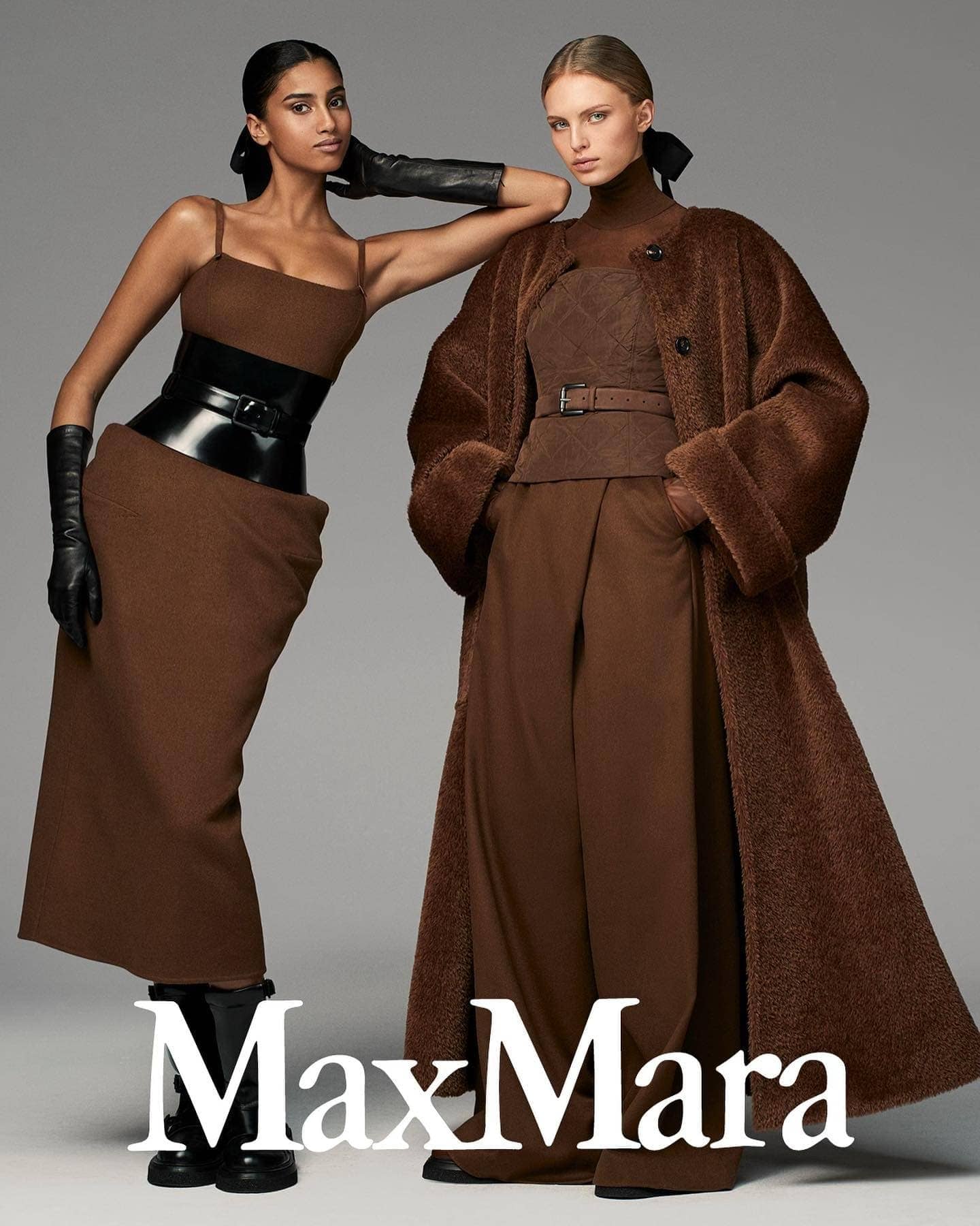 Two fashion models in brown outfits pose in a studio. One wears a wide belt and gloves; the other, a long coat and matching belt.