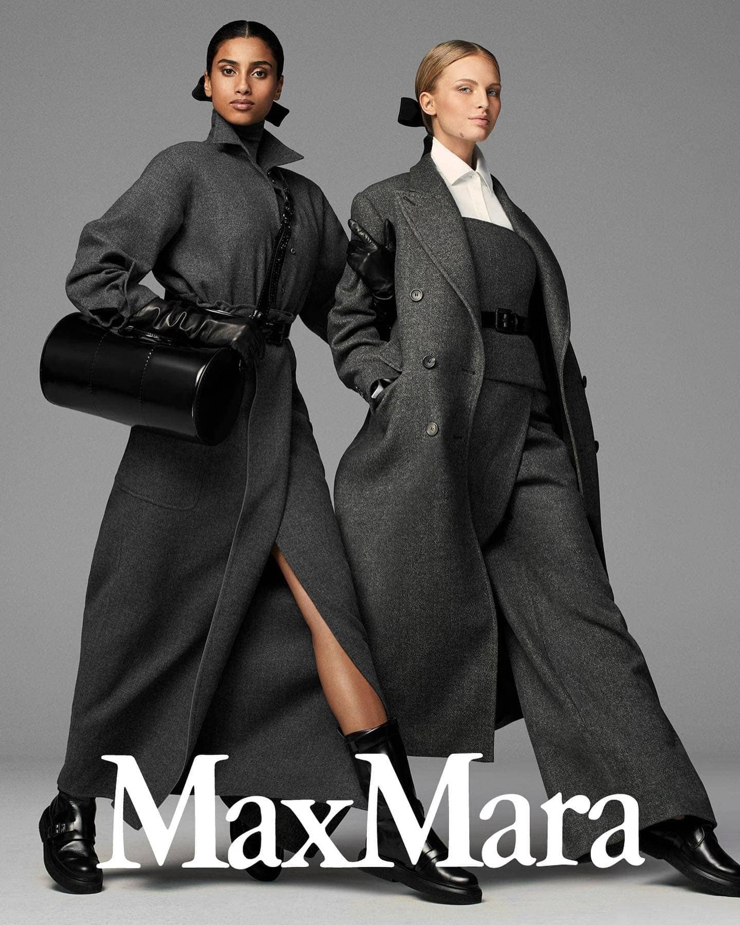 Two fashion models showcase Max Mara coats in a studio shot with a grey background, both wearing black shoes and fashionable grey outerwear.