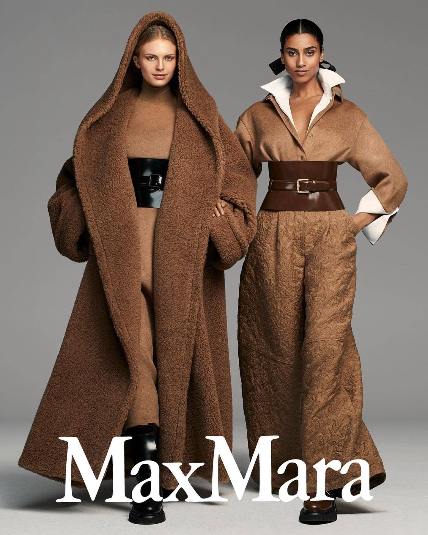 Two fashion models showcase Max Mara apparel in a studio shot featuring long coats, trousers, and belts in matching neutral colors.