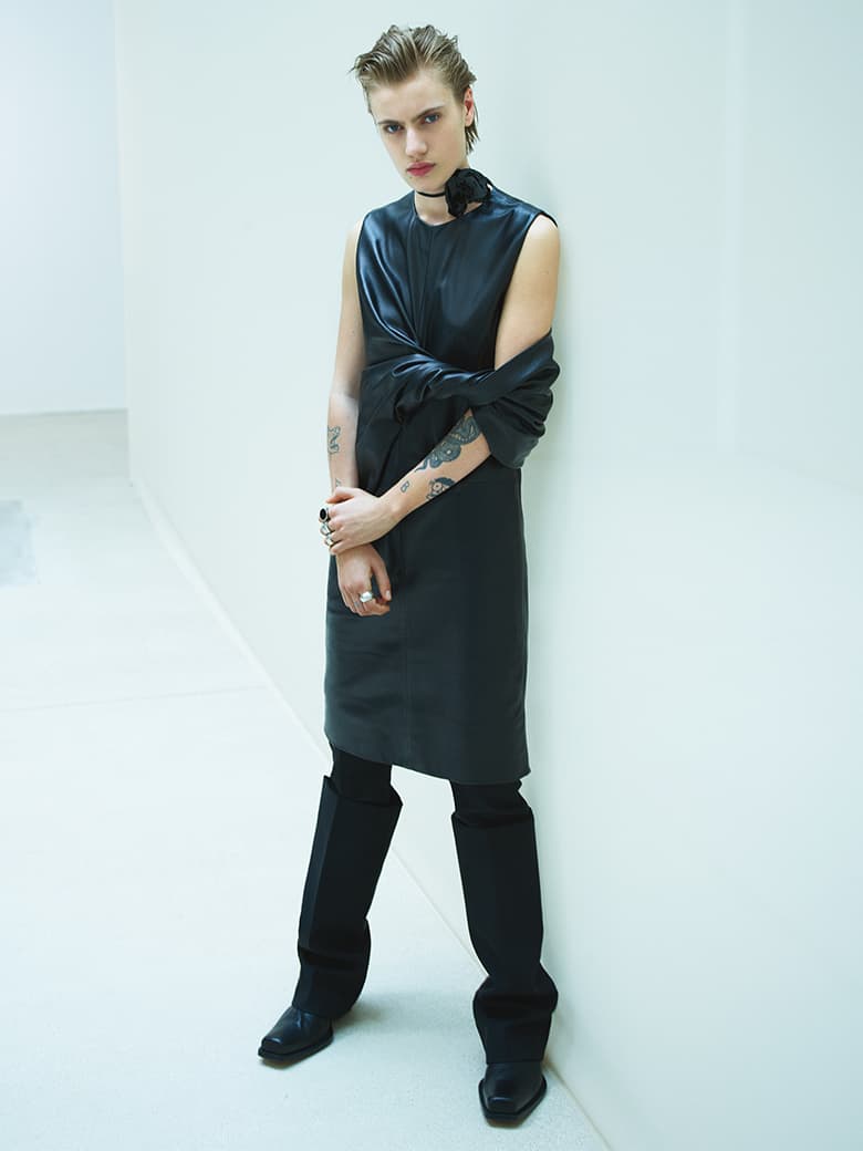 A person with short hair and visible arm tattoos wearing a black sleeveless leather dress and knee-high boots against a white wall.