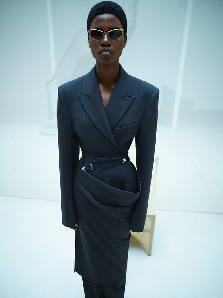 Fashionable dark-skinned adult female posing in a tailored black blazer and skirt ensemble with gold-rimmed sunglasses in a white setting.