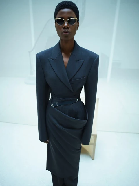 Fashionable dark-skinned adult female posing in a tailored black blazer and skirt ensemble with gold-rimmed sunglasses in a white setting.