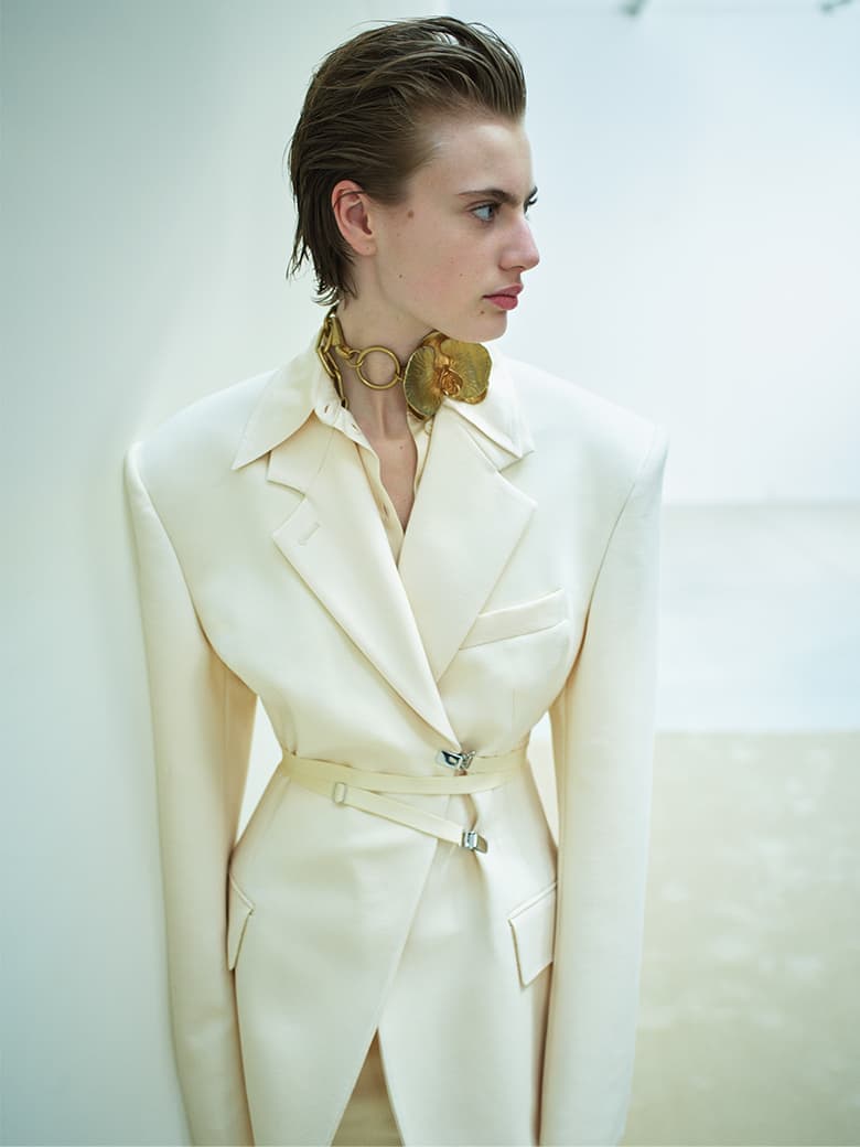 Fashion portrait of a young adult woman in a light yellow suit with an ornate necklace, posing in front of a white wall.