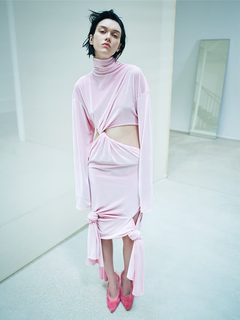 A model poses in a light pink, long-sleeved dress with cutouts, paired with matching heels, against a minimalist white backdrop in a studio setting.