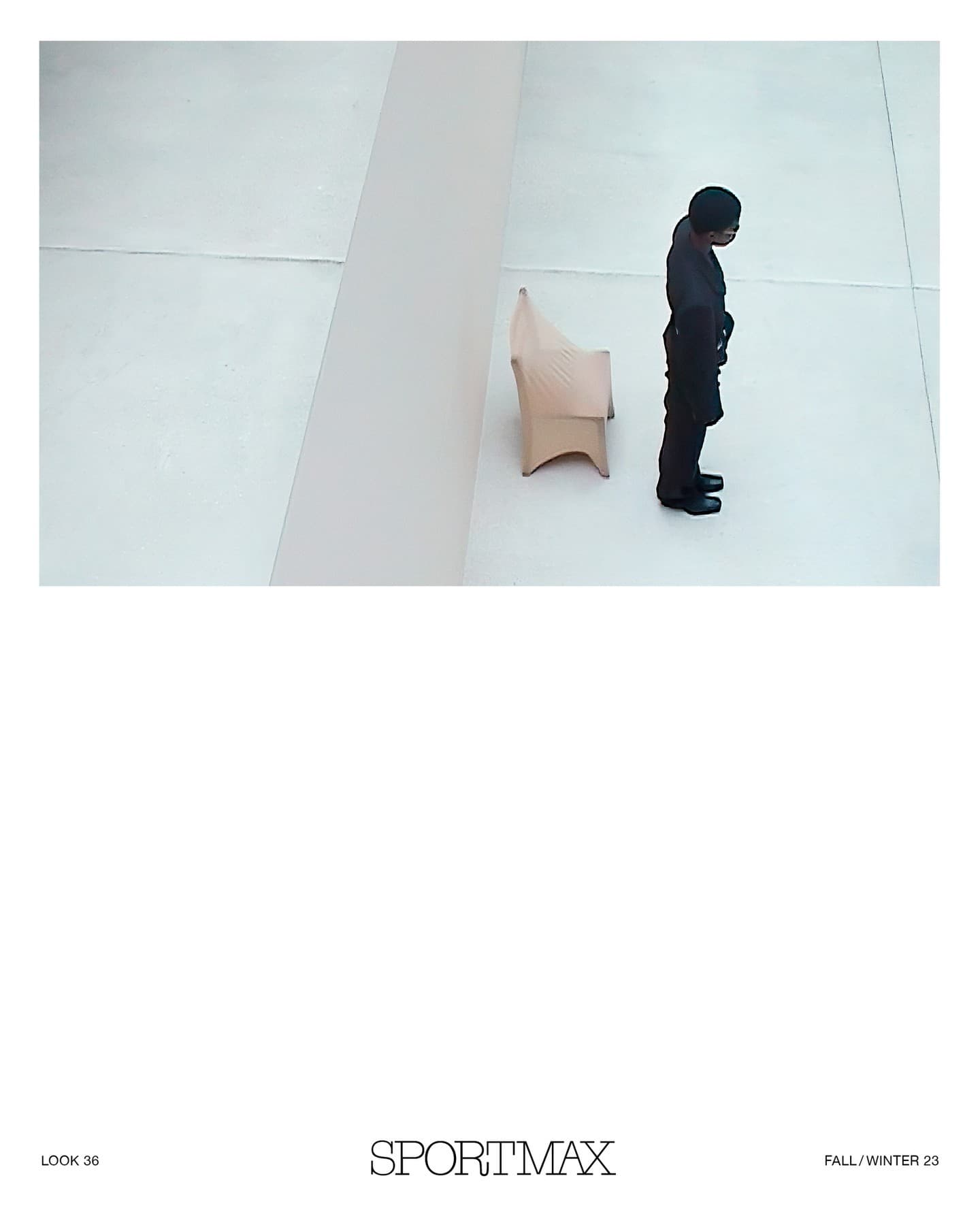 High-angle shot of an adult wearing a black suit standing next to a beige modern chair against a plain white wall in a studio.