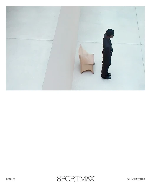 High-angle shot of an adult wearing a black suit standing next to a beige modern chair against a plain white wall in a studio.