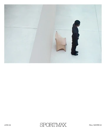 High-angle shot of an adult wearing a black suit standing next to a beige modern chair against a plain white wall in a studio.