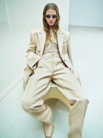 Stylish woman in beige pantsuit and sunglasses poses indoors against a white wall, exuding modern fashion and minimalist elegance.