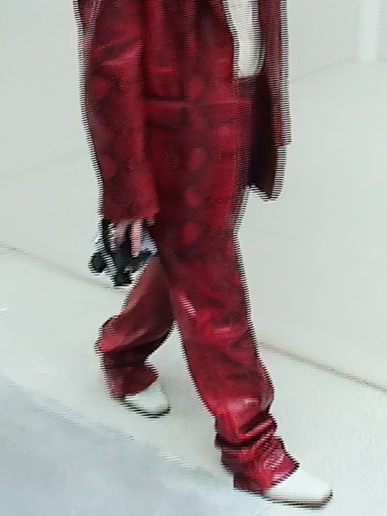 Runway shot of a person wearing a patterned red suit with white dress shoes walking on a white floor. Fashion and style.