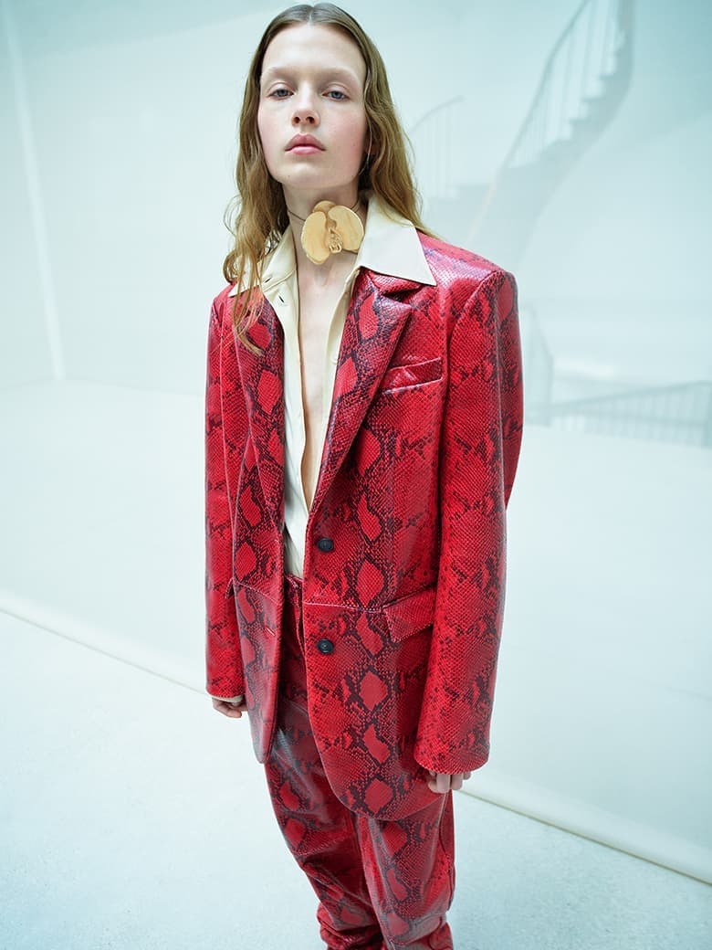 Adult fashion model in red snakeskin-print suit stands in a stark studio with architectural staircase visible, shot in portrait orientation.