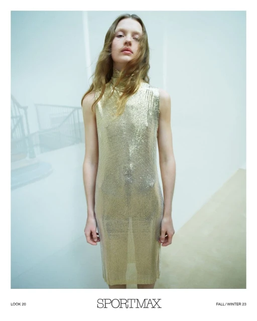 A fair-skinned woman with wavy light brown hair poses indoors wearing a sparkling gold sleeveless midi dress and neutral makeup.
