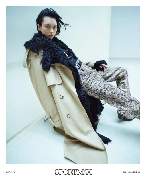 Fashion shot of an adult woman in snake-print pants, a black top, and a beige coat with fur trim, posing in front of a white wall.