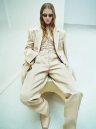 Model wearing oversized beige suit and sunglasses seated against a white background