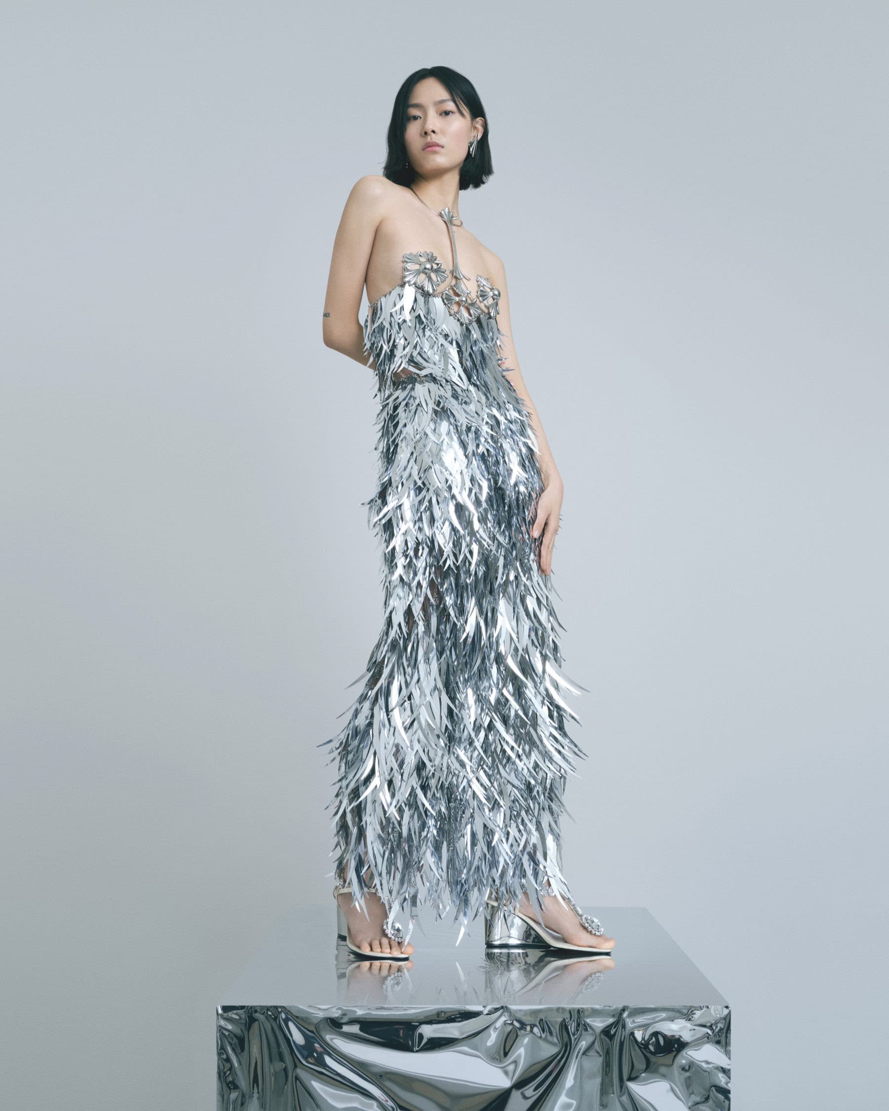 Elegant Asian woman in silver feather dress stands on reflective platform in a fashion portrait with a minimalist, neutral backdrop.