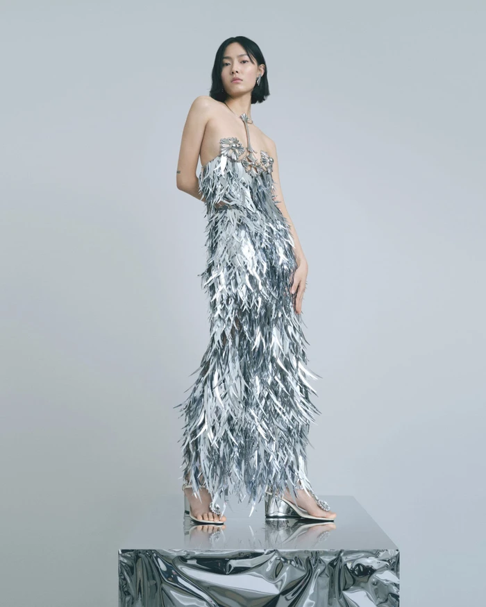 Model Jiali Zhao poses in a metallic silver fringed strapless gown atop a reflective pedestal against a grey studio backdrop.