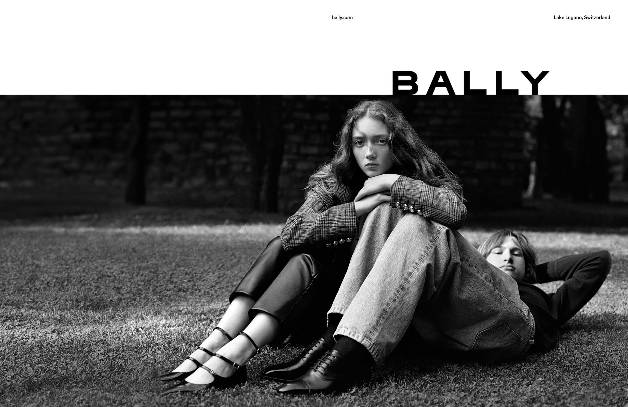 Black and white portrait of a young woman in a blazer and a man in jeans relaxing on the grass with the Bally logo displayed above.