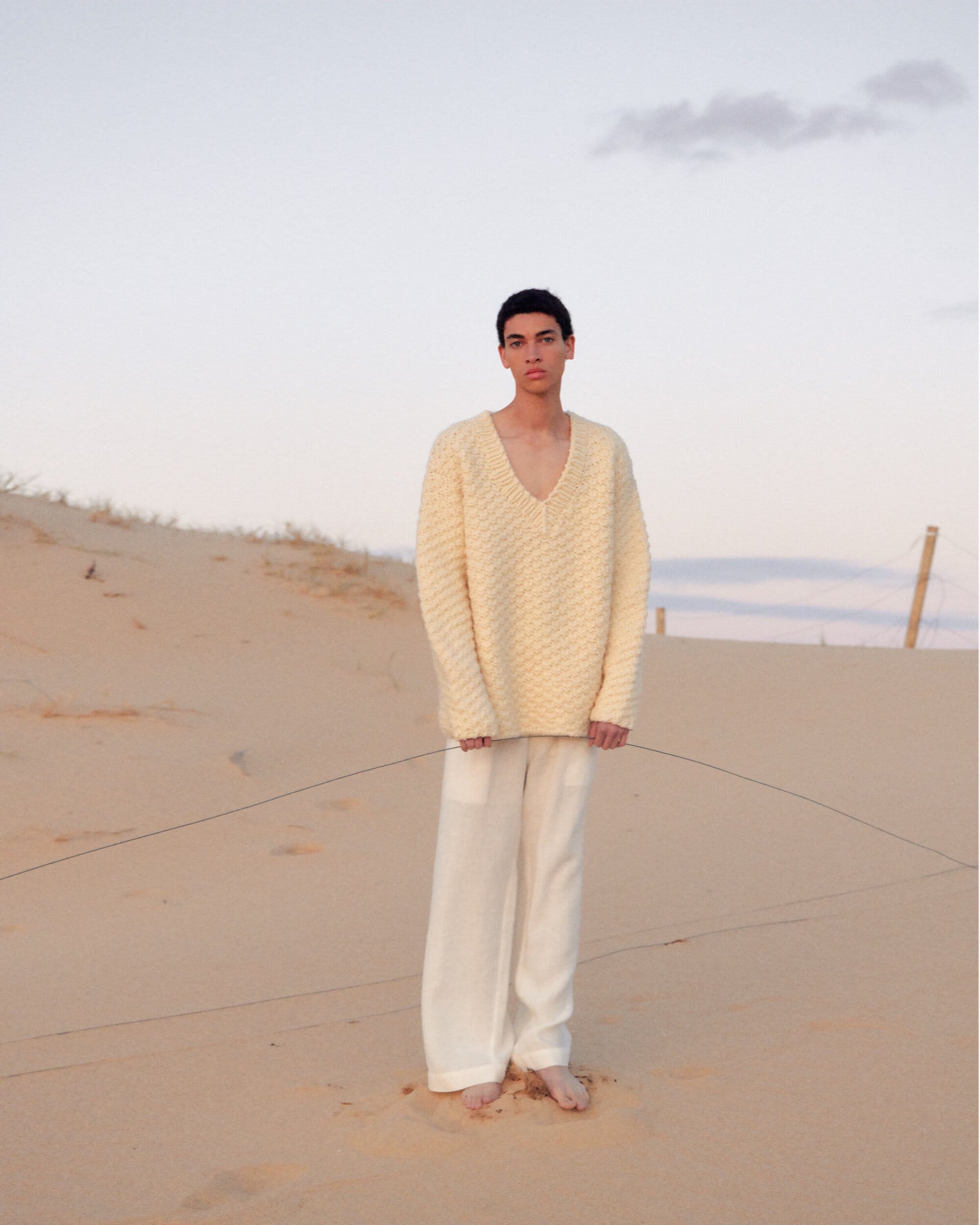 Man stands on sand dune in a cream-colored sweater and white pants. The sky is light blue and the sand is a light golden color.