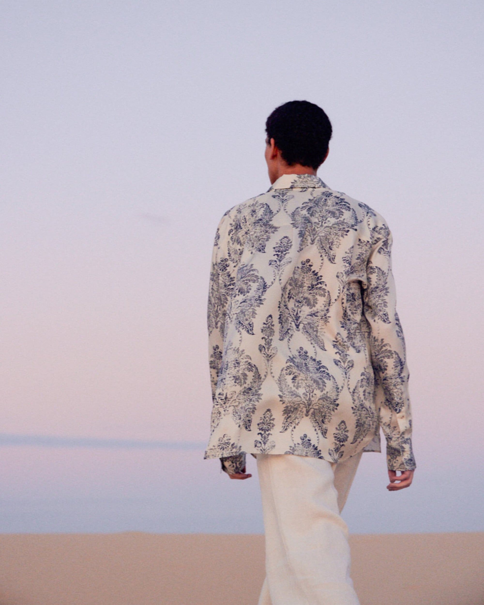 Back view of adult male wearing patterned shirt walking on sand in an outdoor setting against a gradient pink and blue sky.